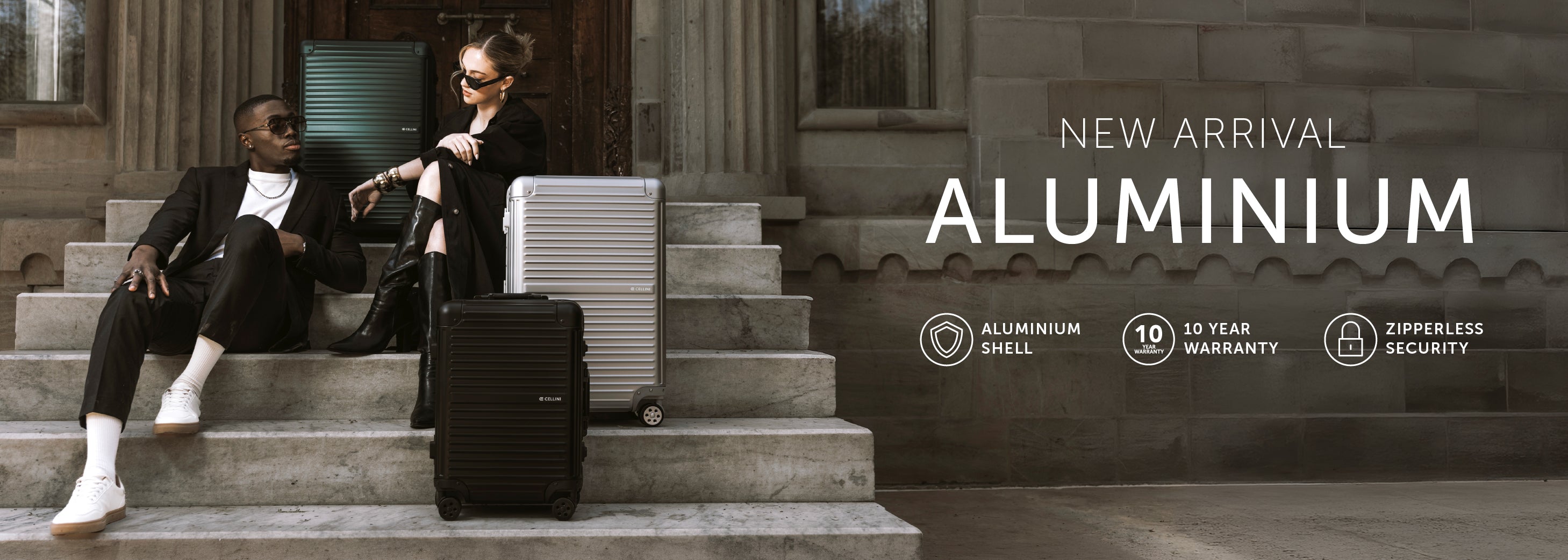 Two models in black suits sit on stone steps beside silver and black hard-shell suitcases, with large text reading 'NEW ARRIVAL ALUMINIUM' and feature icons on the right.